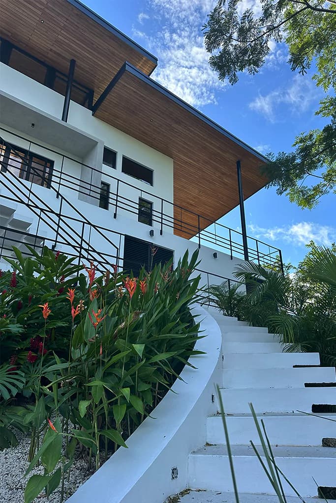White hillside home with wood eaves and a curved stairway through tropical gardens